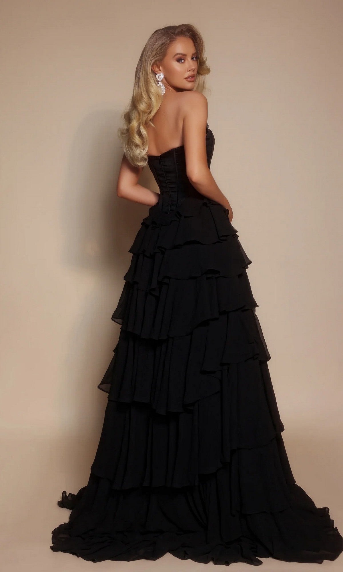 A woman with long blonde hair poses against a beige background in the Portia and Scarlett PS26024 long prom dress, featuring a strapless velvet corset bodice and tiered ruffle skirt, elegantly looking over her shoulder.